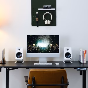 Bàn đứng đen với iMac, hai loa bookshelf trắng, MacBook đứng, sách, ghế da và pegboard xanh đậm treo tường