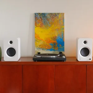 Dàn âm thanh Kanto OBI3 đen giữa hai loa bookshelf Kanto REN trắng trên tủ gỗ, tranh trừu tượng phía sau