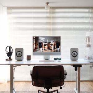 Góc làm việc sáng với bàn đứng trắng, màn hình lớn, cặp loa KEF LSX II LT, tai nghe Marshall trên pegboard, ghế da nâu và cửa sổ rèm