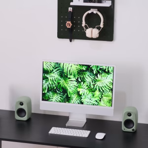 Góc làm việc tối giản với iMac wallpaper lá nhiệt đới, hai loa desktop xanh bạc hà, bàn phím chuột Apple và pegboard xanh đậm treo tường