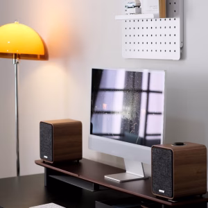 iMac giữa hai loa bookshelf vân gỗ trên kệ nâng màn hình, đèn sàn vòm cam, pegboard trắng treo tường