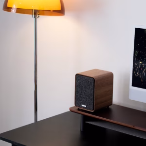 Loa bookshelf Ruark Audio vân gỗ và lưới vải xám trên kệ gỗ, đèn sàn cam và iMac phía sau