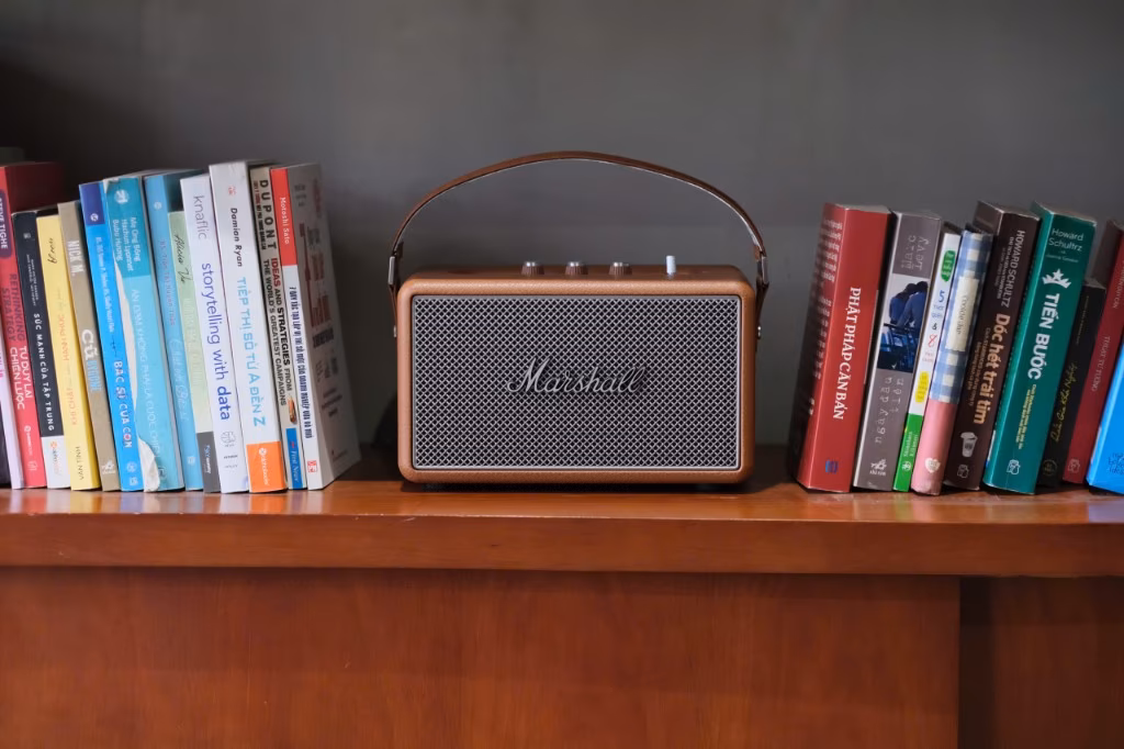 Loa Marshall Kilburn II trên kệ gỗ tối màu giữa hai hàng sách tiếng Việt, tường xám đậm