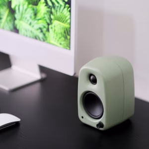 Loa nhỏ màu xanh bạc hà trên bàn đen, phía sau là iMac màn hình lá cây nhiệt đới