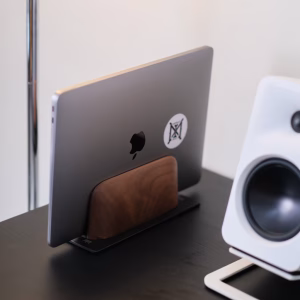 MacBook đóng đứng trong giá laptop gỗ óc chó và kim loại, cạnh loa desktop trắng trên bàn đen