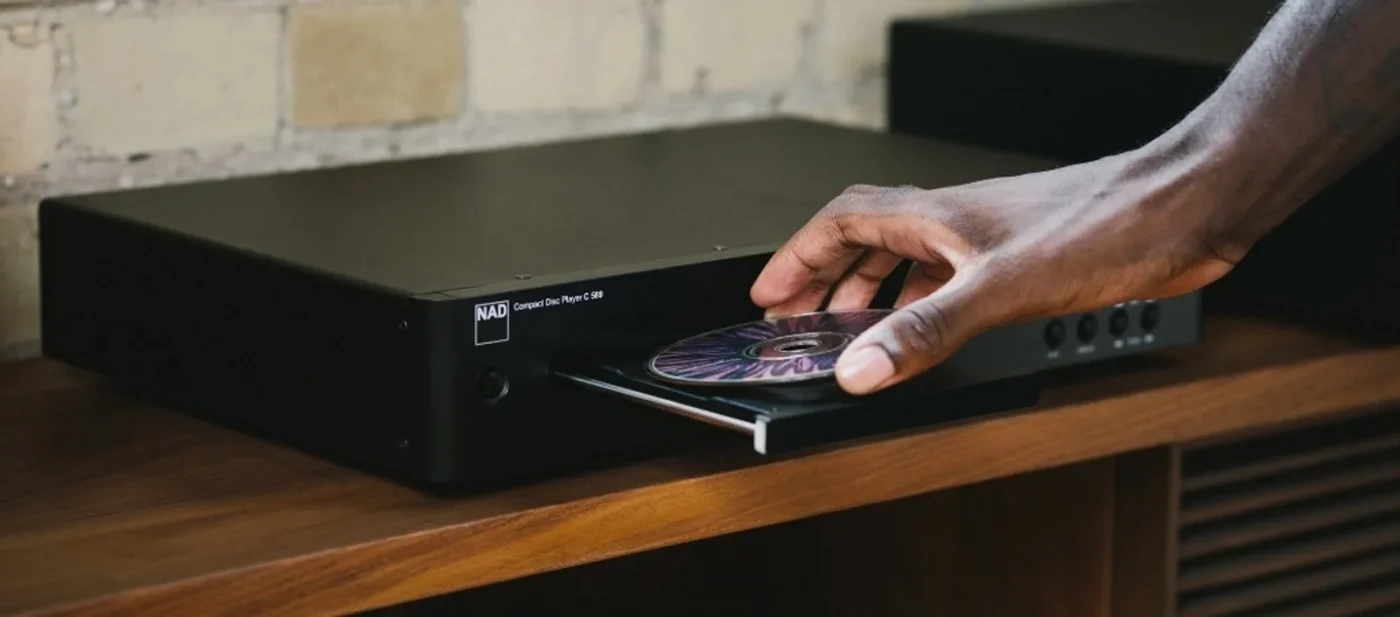 Tay đặt đĩa CD lên khay máy NAD Compact Disc Player trên kệ gỗ, nền tường gạch