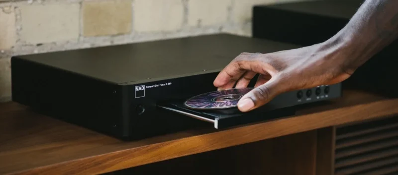 Tay đặt đĩa CD lên khay máy NAD Compact Disc Player trên kệ gỗ, nền tường gạch