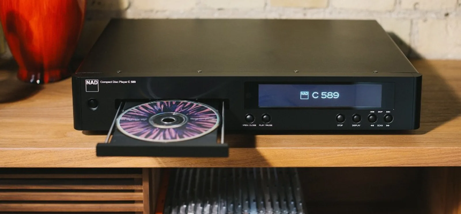 Đầu CD NAD C 589 màu đen mờ, khay đĩa mở với CD màu, màn hình hiển thị NAD C 589 trên kệ gỗ sáng
