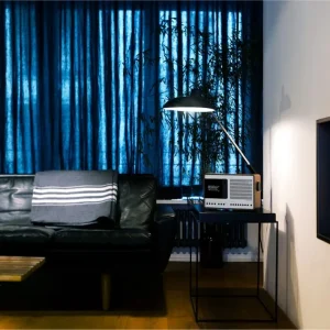 Revo SuperConnect trên bàn metal đen cạnh sofa da đen, rèm xanh và đèn sàn trong phòng khách hiện đại.