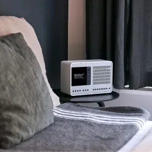 Radio Revo SuperConnect màu trắng trên bàn đầu giường, phòng ngủ tối giản với gối và chăn xám.