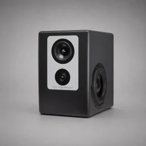 Barefoot Footprint02 góc ba phần tư: mặt trước plate bạc với mid, tweeter, LED xanh và chữ BAREFOOT; cạnh phải woofer bắn ngang với surround cao su; thùng đen mờ, nền xám trung tính, bóng đổ nhẹ dưới đế