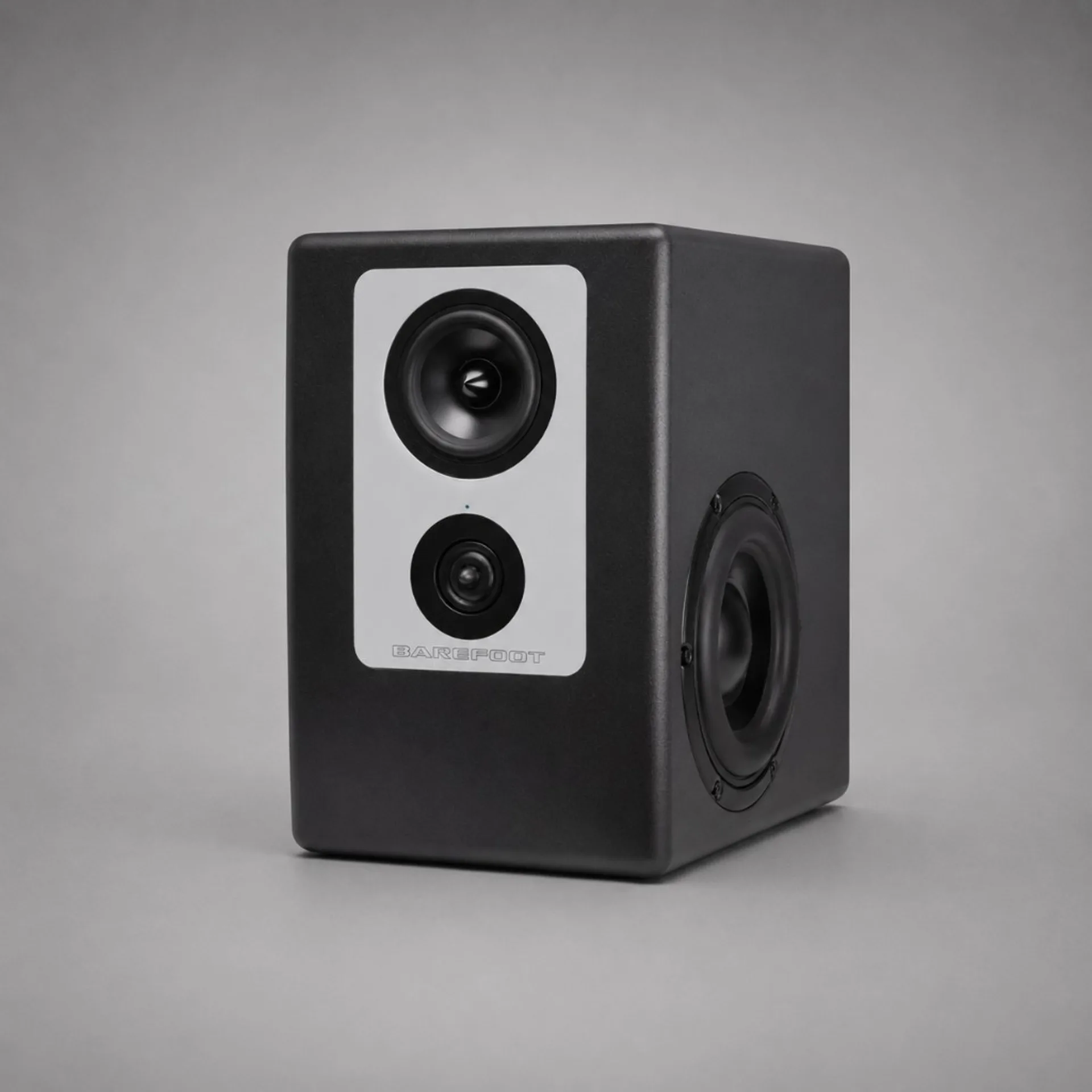 Barefoot Footprint02 góc ba phần tư: mặt trước plate bạc với mid, tweeter, LED xanh và chữ BAREFOOT; cạnh phải woofer bắn ngang với surround cao su; thùng đen mờ, nền xám trung tính, bóng đổ nhẹ dưới đế