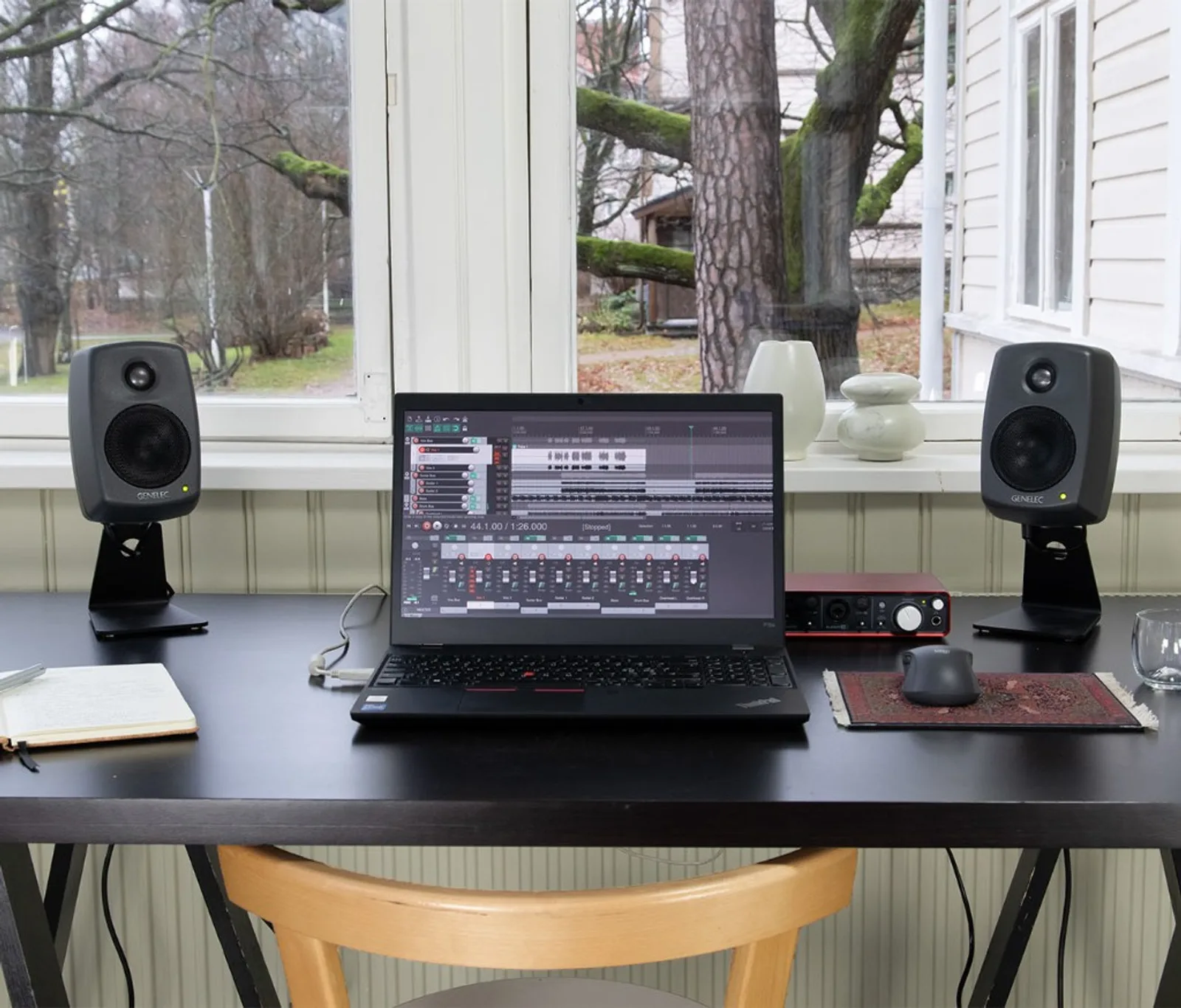 Hai Genelec 8010A xám trên chân L đen hai bên laptop ThinkPad mở DAW; interface đỏ, chuột không dây trên pad thảm, cửa sổ sáng phía sau — không gian làm nhạc tại nhà
