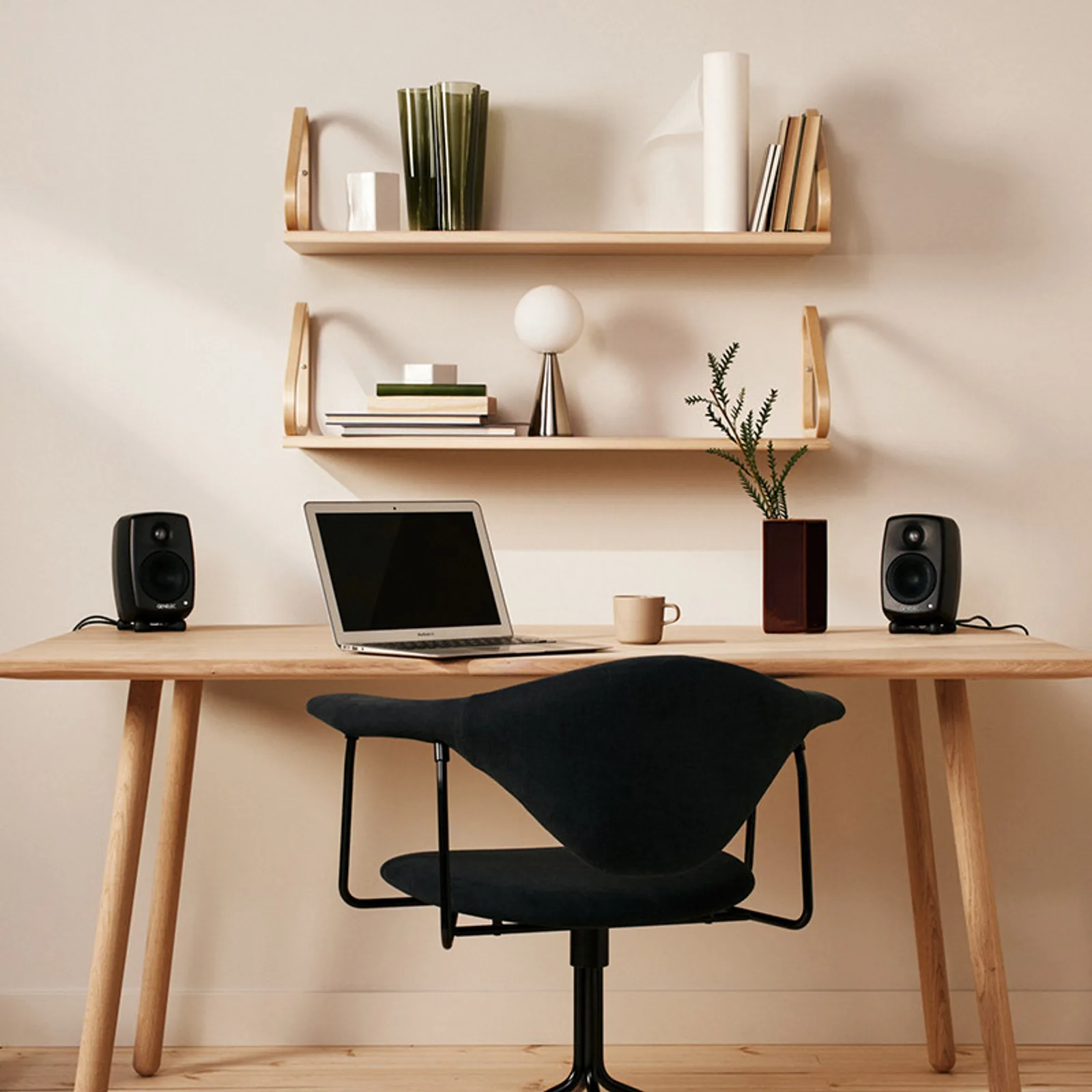Hai Genelec G One đen hai bên laptop MacBook trên bàn gỗ; ghế công thái học đen, kệ treo tường, bình cây — không gian làm việc tối giản Bắc Âu