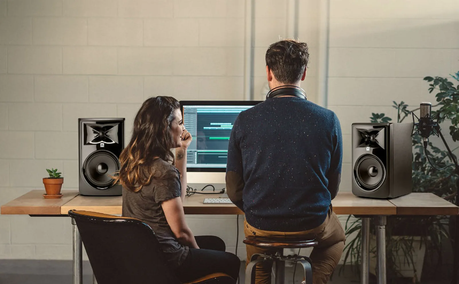 Không gian phòng thu với cặp loa JBL 306P MkII hai bên màn hình DAW, micro condenser, bàn gỗ và tường gạch trắng; hai người làm việc cùng nhau.