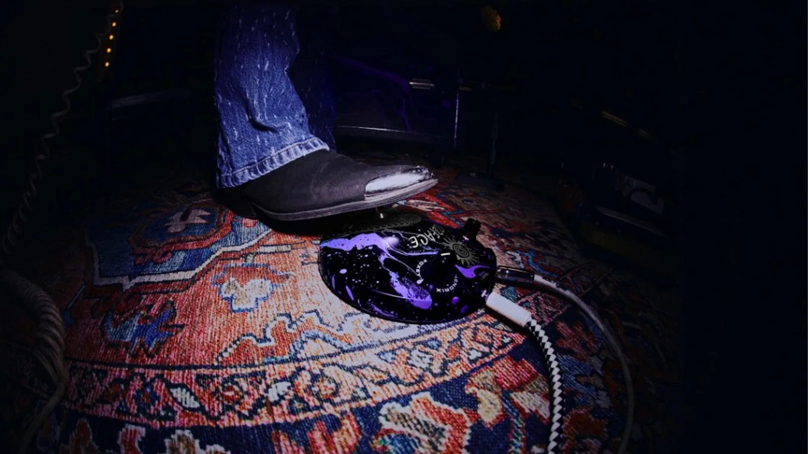 Góc máy fisheye: pedal tròn Fuzz Face họa tiết tím-đen vũ trụ, chữ FUZZ FACE và HENDRIX, hai núm đen, dây jack bọc sọc; bàn chân giày da đen mũi kim loại trên thảm hoa văn, ánh sáng spotlight