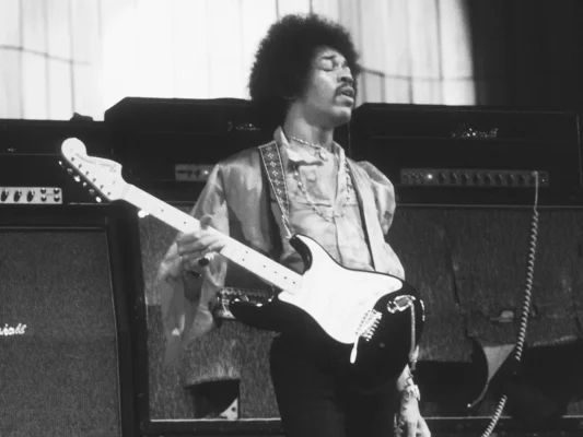 Ảnh đen trắng: Jimi Hendrix chơi Fender Stratocaster ngược tay, mắt nhắm, phía sau là chồng ampli Marshall với logo script nổi bật — khoảnh khắc biểu diễn huyền thoại