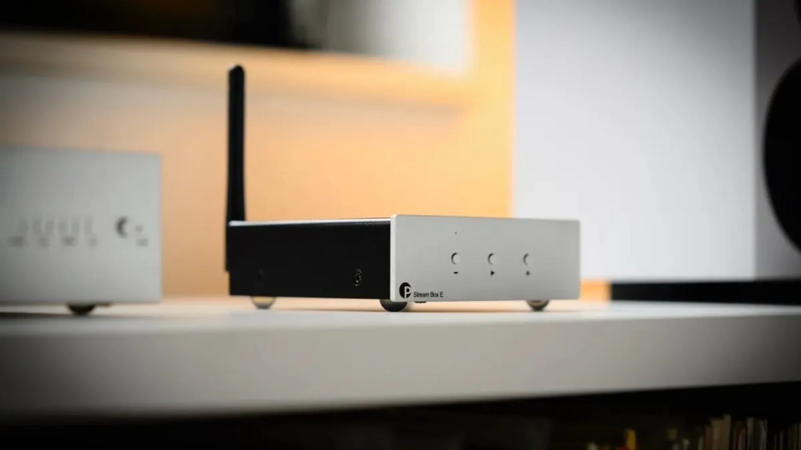 Pro-Ject Stream Box E đặt trên kệ trắng: mặt trước kim loại bạc với logo P, chữ Stream Box E, ba nút tròn (-, play/pause, +), anten đen phía sau, thân đen và chân bạc