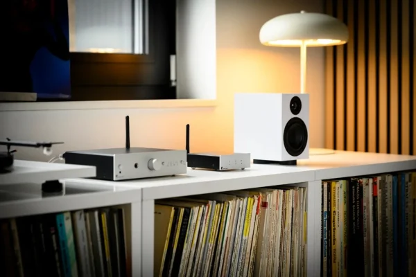 Bối cảnh phòng nghe: Pro-Ject Stream Box E (mặt bạc, volume, tai nghe) và Wireless Box E nhỏ hơn cùng anten, đặt cạnh loa bookshelf trắng, mâm đĩa than trắng và đèn bàn trên kệ trắng chứa đĩa than