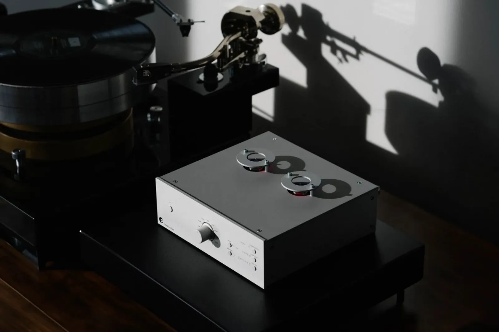 Pro-Ject Tube Box DS3 B màu bạc đặt cạnh mâm đĩa than trong không gian tối, ánh sáng đổ bóng mạnh