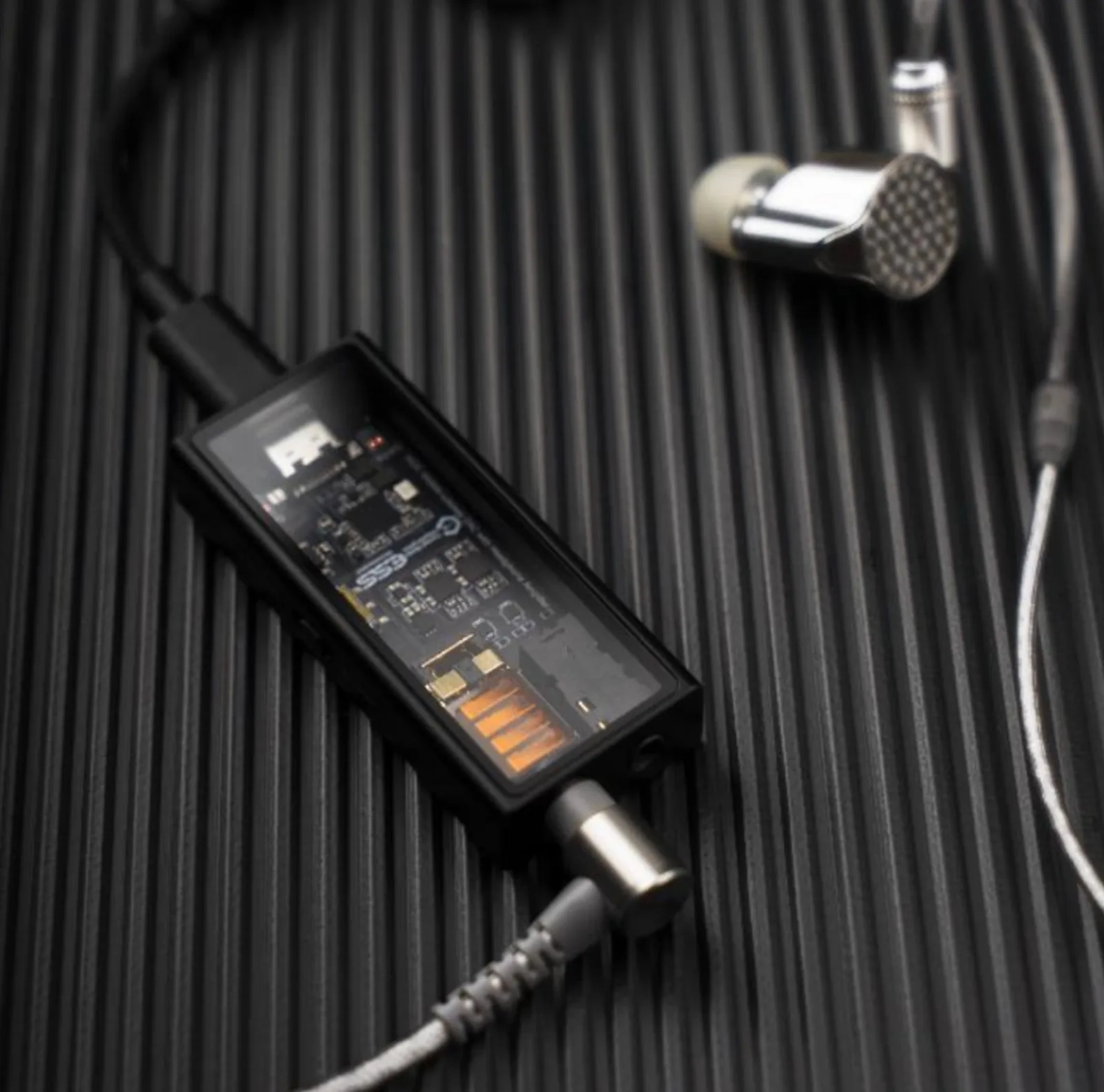 Questyle M15i mặt trong suốt lộ chip ESS và tụ cam, nối USB-C và tai nghe in-ear kim loại dây bạc trên nền đen có vân dọc.